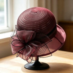 Puede incluir: Un sombrero tipo cloche de color burdeos intenso con una corona estructurada en espiral y un ala ancha semitransparente. El sombrero está adornado con una gran flor de organza superpuesta en un tono burdeos a juego y detalles de red negra.
