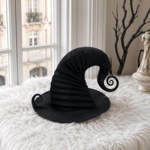 May include: A black witch hat featuring a wide, flat brim and a tall, curved, pointed crown. The fabric of the hat has a subtle horizontal ribbed texture, and the tip of the crown curls downward into a distinct spiral shape.