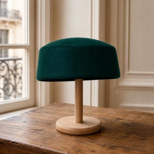 May include: A deep forest green felt hat resting on a light-colored wooden stand. The hat features a flat, circular top and a structured, cylindrical crown. The wooden stand consists of a vertical post attached to a circular base.