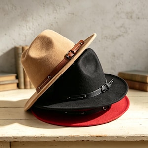 May include: A stack of three fedora hats in tan, black, and red. Each hat features a structured crown, a wide brim, and a decorative band made of faux leather with a small metal buckle and side studs. The hats are layered on top of each other.