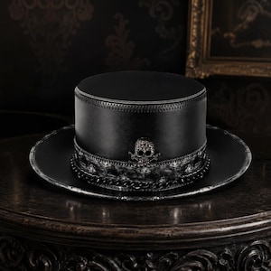 May include: A black top hat featuring a flat crown and a wide brim. The base of the crown is adorned with a decorative band, a metallic chain, and a central skull emblem embellished with small, sparkling clear stones that catch the light.