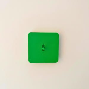 May include: A bright green square wall hook featuring a flat, smooth surface with rounded corners and a single, centred cylindrical peg extending outward from the middle of the square base for hanging items securely on a vertical surface.