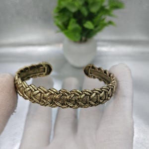 Puede incluir: Un brazalete de metal en tono dorado con un patrón de cuerda trenzada texturizada en la superficie exterior. El brazalete tiene un diseño abierto con un acabado antiguo ligeramente desgastado y detalles visibles en la estructura interior.