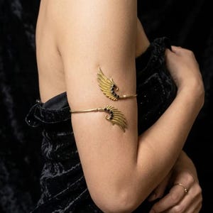 May include: A gold-toned metal arm cuff featuring two symmetrical wing-shaped motifs. Each wing is detailed with textured feathers and accented with three small, round, black stones embedded along the inner curve of the wing structure.