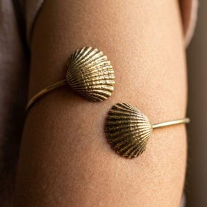 Op de afbeelding: Een goudkleurige open armband met twee identieke, getextureerde schelpmotieven aan de uiteinden. De metalen schelpen hebben een geribbeld oppervlak en zijn bevestigd aan een dunne, gladde, ronde band die om de arm sluit met een opening ertussen.