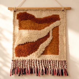 May include: A square wall hanging featuring a tufted design with cream, burnt orange, and rust-coloured yarn. The piece is suspended from a wooden dowel and finished with a bottom row of braided fringe tassels in cream, black, and pink yarn.