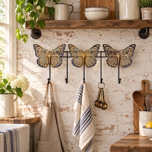 May include: A black metal wall-mounted rack featuring three decorative butterfly ornaments with gold and blue distressed finishes. The rack includes five hooks for hanging items, with a beige apron, a blue and white striped towel, and measuring spoons.