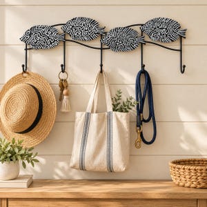 May include: A black metal wall-mounted hook rack featuring four decorative fish silhouettes with white maze-like patterns. The rack has five hooks holding a straw hat, a keychain with a tassel, a beige canvas tote bag, and a navy blue rope leash.
