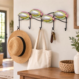 May include: A black metal wall-mounted hook rack featuring four decorative fish figures with silver, yellow, and pink painted details. The rack has five hooks, with a straw hat, a plain canvas tote bag, and a set of keys hanging from them.