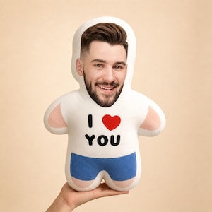 May include: A white plush doll shaped like a person with a photographic print of a smiling person's face on the head. The body features the text I LOVE YOU with a red heart icon and blue shorts printed on the front of the white fabric surface.