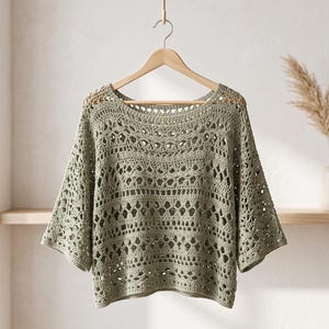 Op de afbeelding: Een saliegroene gehaakte top met een brede boothals, driekwartmouwen en een ingewikkeld opengewerkt patroon met herhalende geometrische en bloemmotieven over het hele kledingstuk, gepresenteerd op een houten kledinghanger.