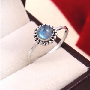 Könnte beinhalten: Ein Silberring mit einem runden, irisierenden Mondstein-Cabochon in einer Fassung. Der Stein ist von einem dekorativen Rand aus kleinen, erhabenen Silberkugeln umgeben. Der Ring ist schmal und poliert und weist eine glatte, metallische Oberfläche auf.