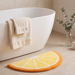 Puede incluir: Una alfombrilla de baño semicircular con forma de rodaja de limón, con segmentos amarillos, bordes blancos y un borde exterior naranja. Una toalla beige doblada cuelga de una bañera blanca, junto a un jarrón de cristal con ramas de eucalipto.