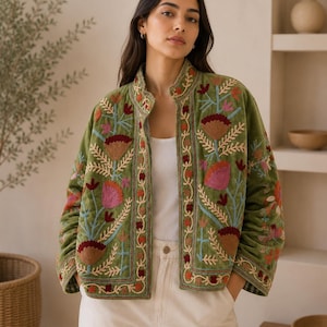Può includere: Una giacca in velluto verde oliva con colletto alto e apertura frontale. La giacca è decorata con intricati ricami floreali nei toni del rosa, bordeaux e azzurro, con maniche lunghe e un orlo dritto sopra un top bianco.