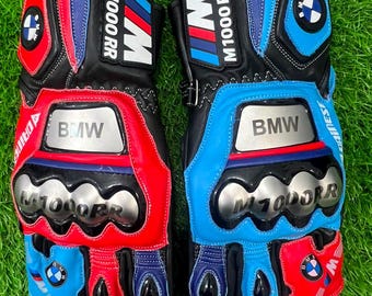 Puede incluir: Un par de guantes de carreras para motocicleta con paneles de cuero negro, rojo y azul. Los guantes muestran el logotipo de BMW, el texto M1000RR y protectores de nudillos plateados. El diseño incluye detalles cosidos y marcas.