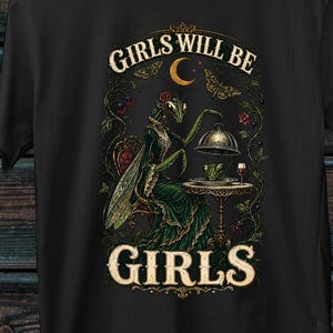 Op de afbeelding: Een zwart t-shirt met korte mouwen en een grafische print van een bidsprinkhaan in een groene jurk, zittend aan een tafel met een stolp en een glas. De tekst "GIRLS WILL BE GIRLS" is in een crèmekleurig lettertype boven en onder de afbeelding gedrukt.