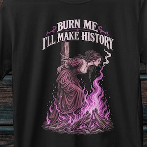 Può includere: Una t-shirt nera a maniche corte con una stampa grafica che raffigura una persona legata a un palo di legno con fiamme viola che si alzano da un fuoco sottostante. Il testo sopra la figura recita "BURN ME I'LL MAKE HISTORY" in bianco.