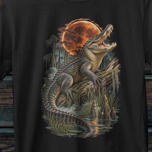 Op de afbeelding: Een zwart t-shirt met korte mouwen en een grafische print van een alligator op een boomstam in een moerasachtige omgeving onder een grote, gloeiende oranje maan. Het ontwerp toont gedetailleerde texturen van de schubben en de vegetatie.