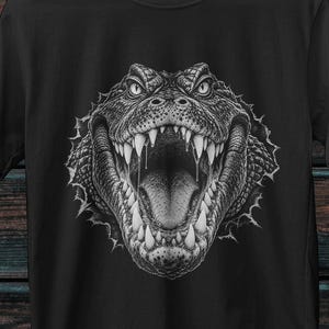 Może przedstawiać: Czarny t-shirt z krótkim rękawem z detaliczną, monochromatyczną grafiką głowy aligatora z szeroko otwartą paszczą, ukazującą ostre zęby. Wzór wygląda, jakby wyłaniał się z poszarpanego, okrągłego rozdarcia w materiale koszulki.