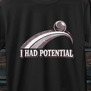 Puede incluir: Una camiseta negra de manga corta con un gráfico de una esfera rodando sobre un arco curvo. Debajo del gráfico, el texto "I HAD POTENTIAL" aparece en letras mayúsculas blancas. La camiseta tiene un cuello redondo y un diseño estándar.
