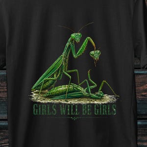 Op de afbeelding: Een zwart t-shirt met korte mouwen en een grafische illustratie van twee groene bidsprinkhanen. De ene staat boven de andere en houdt een losgekoppelde kop vast. De tekst "GIRLS WILL BE GIRLS" is in het groen onder de insecten gedrukt.