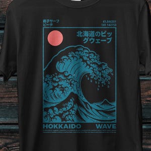 Peut inclure: Un t-shirt noir à manches courtes avec une impression graphique représentant une grande vague bleue, un soleil rose et du texte japonais. Le motif inclut les mots HOKKAIDO WAVE, des coordonnées et des caractères japonais dans un cadre.