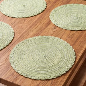 May include: A set of five round, light green placemats featuring a coiled, braided texture. Each placemat is constructed from woven material with a consistent circular pattern, radiating from the center to a decorative braided edge finish.