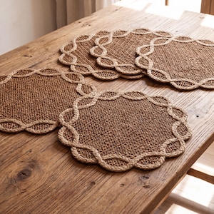 May include: A set of three round placemats made of woven brown natural fiber. Each placemat features a textured, tightly woven center and a decorative border consisting of a braided, looped rope design that creates a scalloped edge effect.