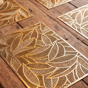 May include: A set of four rectangular gold-coloured placemats featuring a detailed laser-cut pattern of overlapping leaves. Each placemat has a metallic finish and a consistent design of veins and foliage, creating a uniform appearance across all pieces.