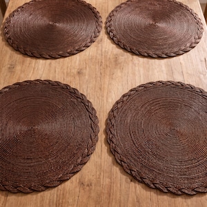 May include: A set of four circular dark brown placemats arranged in a two-by-two grid. Each placemat features a tightly coiled center design with a decorative braided border edge, creating a uniform and textured appearance across all four pieces.