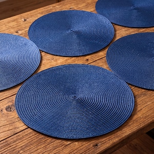 May include: A set of four circular, deep blue placemats featuring a tightly woven, concentric spiral texture. Each placemat is constructed from a durable, braided material that creates a uniform, ridged surface pattern radiating from the centre.