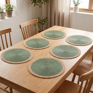 Puede incluir: Un juego de cinco manteles individuales redondos dispuestos sobre una mesa de madera. Cada mantel presenta un centro circular de color verde salvia hecho de material enrollado, rodeado por un borde ancho y texturizado de fibra tejida natural.