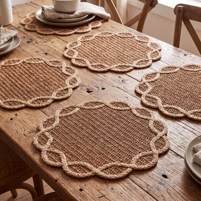 May include: A set of four round, woven placemats made from natural tan fibers. Each placemat features a tight, grid-like center weave surrounded by a decorative, braided rope-style border that creates a scalloped edge around the entire perimeter.