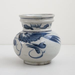 May include: A white ceramic jar with a rounded body and a flared rim, featuring hand-painted blue floral and bird motifs. The glaze has a glossy finish with visible fine crackling throughout the surface, and the base is slightly recessed.