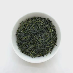 以下が含まれることがあります： 白い陶器のボウルに入った緑茶の茶葉。針のように細い濃いフォレストグリーンの茶葉がぎっしりと詰まっており、滑らかで丸みを帯びた容器の中で質感のある外観を作り出しています。