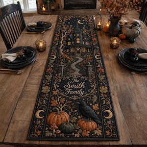 May include: A rectangular black fabric table runner featuring a printed design of a haunted house, a winding path, pumpkins, a black crow, and autumn leaves. The text "The Smith Family" is printed in the centre. The runner has a decorative border.