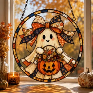 May include: A circular stained glass suncatcher featuring a white ghost with a large patterned bow on its head, holding an orange jack-o'-lantern filled with colourful autumn leaves and berries, suspended by a black metal chain in a window frame.