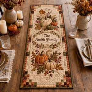 Puede incluir: Un camino de mesa acolchado rectangular de tela color crema con motivos otoñales bordados que incluyen calabazas, hojas y bayas. El centro muestra el texto The Smith Family en una tipografía decorativa rodeado por un borde floral.