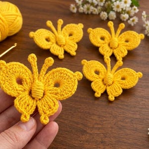 May include: Four bright yellow crocheted butterflies made from yarn, featuring intricate wing patterns and antennae. A matching yellow ball of yarn and a yellow crochet hook are present, alongside small white daisy flowers.