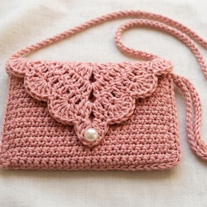 May include: A dusty pink crochet crossbody bag featuring a rectangular body with a scalloped flap closure. The flap is adorned with a single white pearl-like bead at the center. The bag includes a matching crocheted strap for shoulder wear.