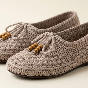 Puede incluir: Un par de zapatillas de ganchillo de color marrón claro con un patrón de punto texturizado, puntera redondeada y un detalle de cordón decorativo en la parte superior de cada zapato adornado con tres pequeñas cuentas de madera.