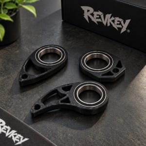 Könnte beinhalten: Drei schwarze, tropfenförmige mechanische Bauteile mit integrierten silberfarbenen Metallkugellagern. Jedes Teil weist eine texturierte, matte Oberfläche und geometrische Ausschnitte auf. Eine schwarze Box mit dem weißen Text REVKEY ist zu sehen.
