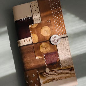 Pode incluir: Uma capa de diário retangular em couro com um design de patchwork de várias peças de couro castanho, bege e creme costuradas com fio branco. Uma etiqueta circular com o texto DALI LEATHER ART está presa por uma banda elástica escura.