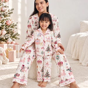 May include: Matching white long-sleeve pajama sets featuring a colorful holiday print with nutcrackers, crowns, Christmas trees, and ballet dancers. The button-down tops have notched collars and pink piping, paired with matching loose-fitting pajama pants.