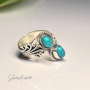 Könnte beinhalten: Ein offener Ring aus Sterlingsilber mit zwei ovalen Türkissteinen mit dunkler Matrix-Maserung. Das Band zeigt komplizierte Schnörkel und gedrehte Kordeldetails um die Fassungen. Der Text SilverAura925 ist in der Ecke aufgedruckt.