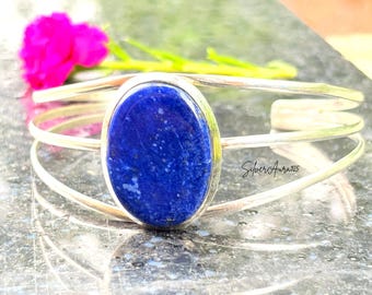 May include: A silver cuff bracelet featuring a large, oval-shaped, deep blue lapis lazuli gemstone set in the centre. The band consists of three thin, polished silver wires that converge at the sides. The text SilverAura925 is visible.
