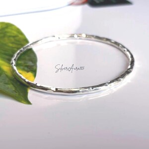Puede incluir: Un brazalete circular y fino de plata con una superficie martillada y texturizada. El metal tiene un acabado pulido y reflectante. El texto "SilverAura925" está impreso en el centro del brazalete, indicando la composición del material.