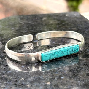 Op de afbeelding: Een zilveren armband met een rechthoekige, helder turquoise steen met donkere aders. De metalen band is glad en gepolijst, met de tekst SilverAura925 gegraveerd op het binnenoppervlak van de armband bij de opening.