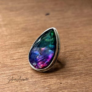 May include: A teardrop-shaped ring featuring a faceted iridescent gemstone with deep blue, purple, and green hues set in a polished silver band. The stone displays a reflective, multi-colored surface with visible internal textures and light refractions.