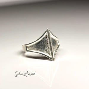 May include: A silver ring featuring a prominent, raised triangular focal point with a textured, matte finish. The band is smooth and polished, tapering towards the back. The text SilverAura925 is printed in black script in the lower left corner.